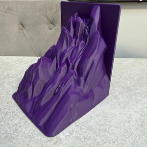 May include: A vibrant purple mountain range bookend. The bookend features a detailed, textured design of a mountain landscape. The item is made of a solid material and is designed to hold books upright on a shelf or desk.