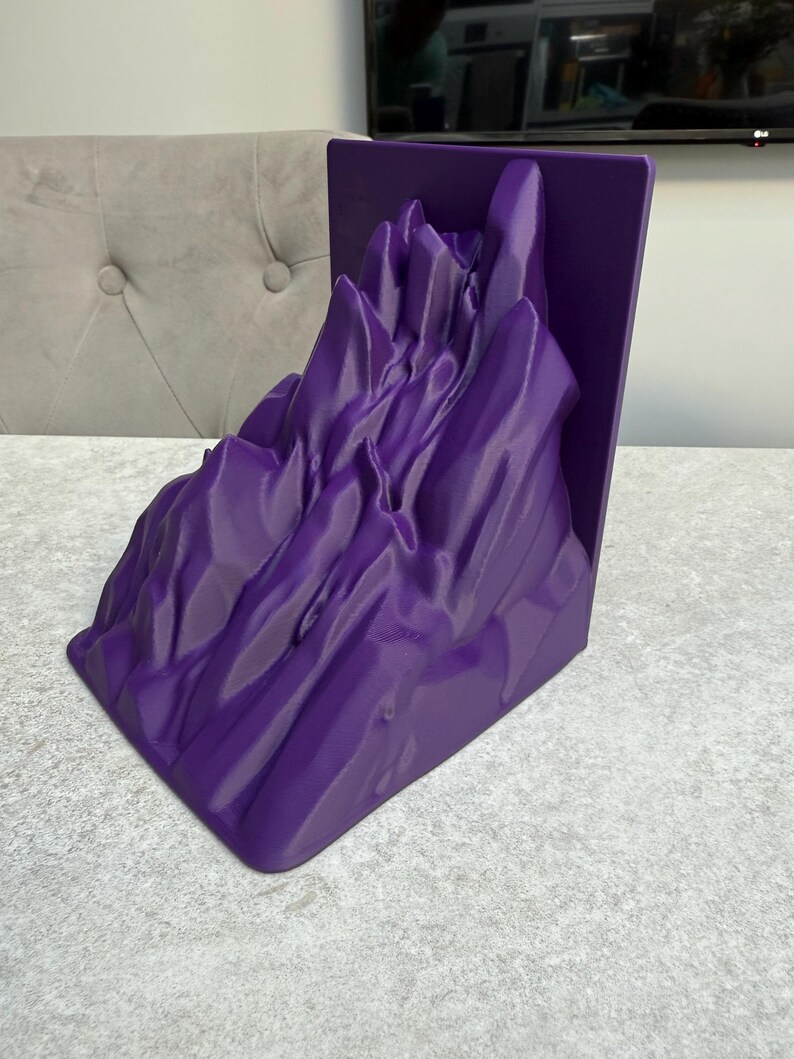 May include: A vibrant purple mountain range bookend. The bookend features a detailed, textured design of a mountain landscape. The item is made of a solid material and is designed to hold books upright on a shelf or desk.