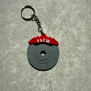 May include: A gray 3D-printed keychain featuring a brake rotor design with a red brake caliper. The red caliper has the text "P & C 3D" in white. The keychain includes a silver key ring and chain.