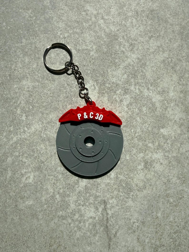 May include: A gray 3D-printed keychain featuring a brake rotor design with a red brake caliper. The red caliper has the text "P & C 3D" in white. The keychain includes a silver key ring and chain.