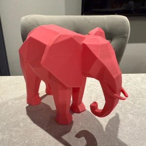 Op de afbeelding: Een koraalkleurige, geometrische olifantsculptuur. Het decoratieve stuk heeft een facetontwerp, waardoor het een moderne, hoekige uitstraling heeft. De olifant staat op vier poten, met een slurf en grote oren.