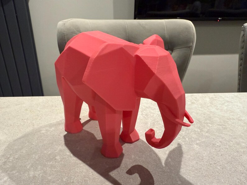 Op de afbeelding: Een koraalkleurige, geometrische olifantsculptuur. Het decoratieve stuk heeft een facetontwerp, waardoor het een moderne, hoekige uitstraling heeft. De olifant staat op vier poten, met een slurf en grote oren.