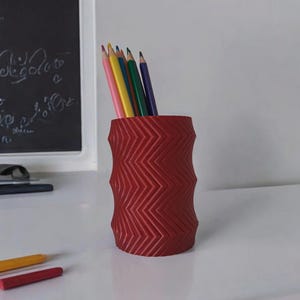 May include: A red pencil holder with a zigzag pattern, holding several colored pencils. The pencils are various colors, including pink, yellow, green, and blue. The holder is on a white surface.