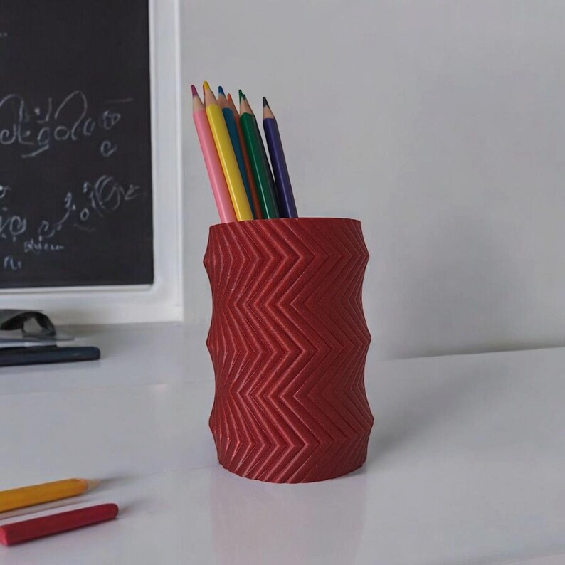 May include: A red pencil holder with a zigzag pattern, holding several colored pencils. The pencils are various colors, including pink, yellow, green, and blue. The holder is on a white surface.