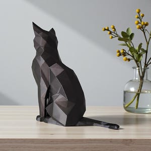 May include: A black geometric cat sculpture sits on a wooden surface. The cat is in a seated position with its tail extended. A clear glass vase with yellow flowers and green leaves is to the right of the cat.