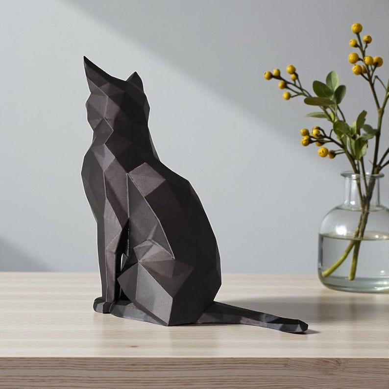 May include: A black geometric cat sculpture sits on a wooden surface. The cat is in a seated position with its tail extended. A clear glass vase with yellow flowers and green leaves is to the right of the cat.