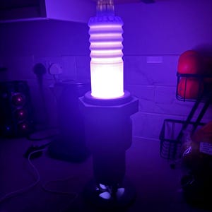 May include: A unique, illuminated lamp shaped like a large spark plug. The lamp has a cylindrical body with horizontal ridges, a hexagonal base, and a top section. The lamp emits a bright, white light, and is set against a dark background.