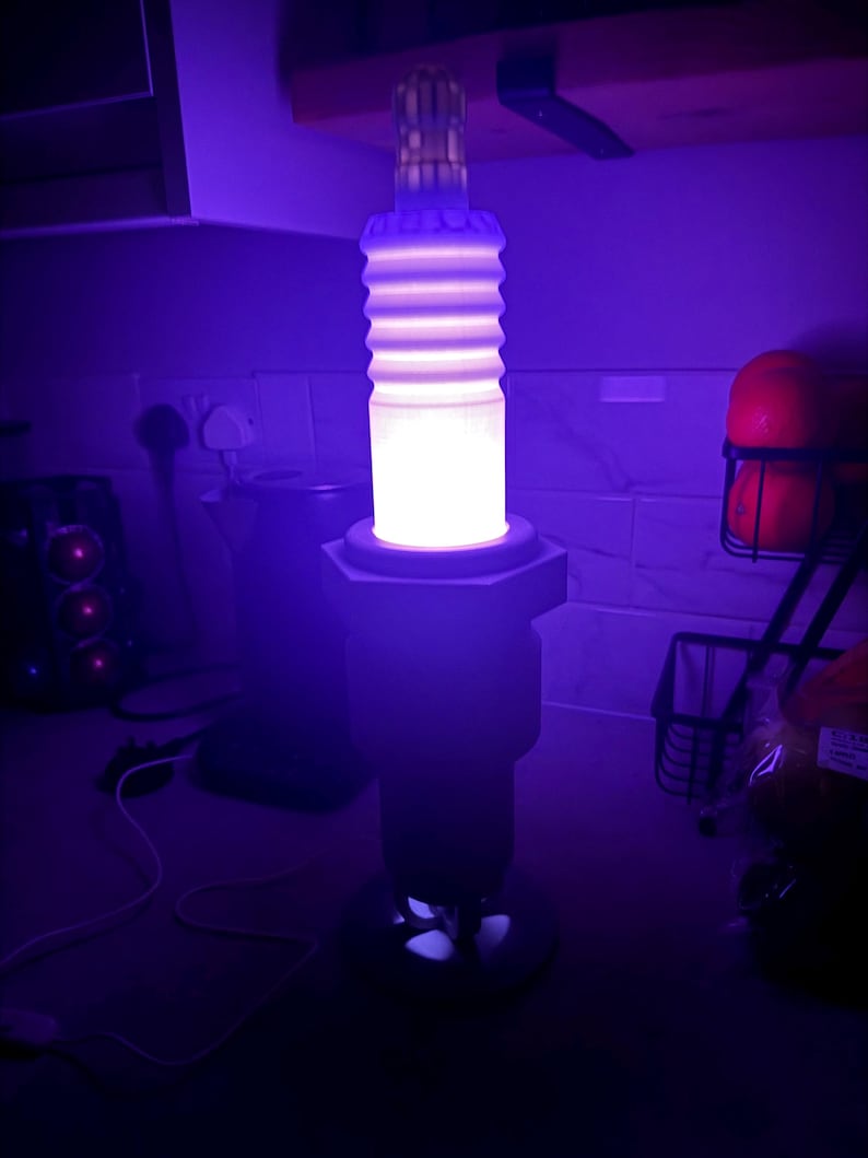 May include: A unique, illuminated lamp shaped like a large spark plug. The lamp has a cylindrical body with horizontal ridges, a hexagonal base, and a top section. The lamp emits a bright, white light, and is set against a dark background.