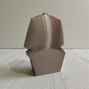 May include: A taupe-colored, geometric sculpture with a stylized, angular design. The sculpture has a rectangular base and a top section with vertical lines. The overall shape resembles a bust or a stylized head.