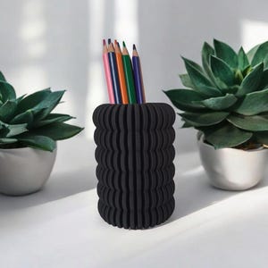 May include: A black, textured pencil holder with a ribbed design, holding several colored pencils. The holder is cylindrical and sits on a white surface, flanked by two potted succulents.