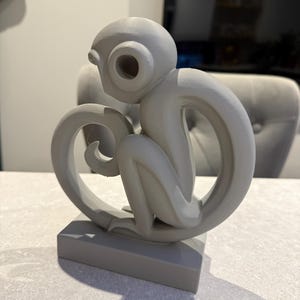 Puede incluir: Una escultura de mono gris con un dise&ntilde;o estilizado. El mono est&aacute; sentado sobre una base rectangular, con su cola formando un bucle. La escultura tiene un acabado liso y mate y est&aacute; hecha de un material s&oacute;lido.