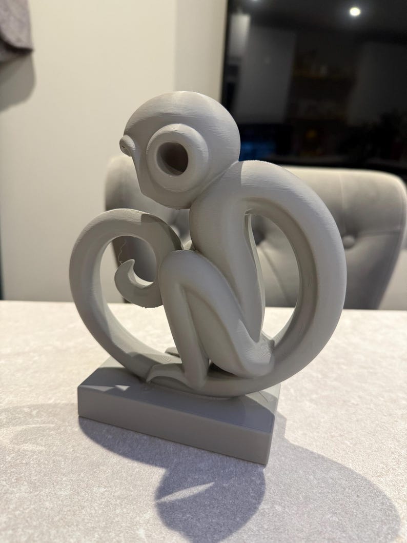 Puede incluir: Una escultura de mono gris con un dise&ntilde;o estilizado. El mono est&aacute; sentado sobre una base rectangular, con su cola formando un bucle. La escultura tiene un acabado liso y mate y est&aacute; hecha de un material s&oacute;lido.