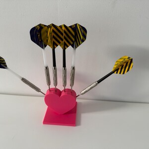 May include: A set of darts with yellow and black striped flights, silver barrels, and black and white shafts, arranged in a pink heart-shaped holder. The darts are displayed against a white background.