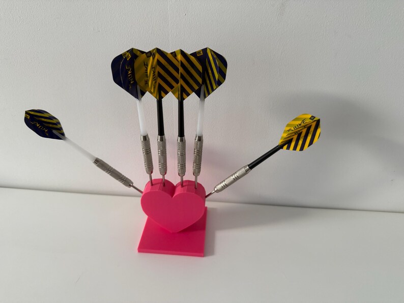 May include: A set of darts with yellow and black striped flights, silver barrels, and black and white shafts, arranged in a pink heart-shaped holder. The darts are displayed against a white background.