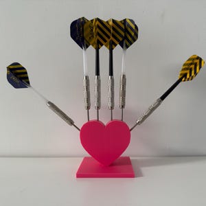 May include: A pink heart-shaped dart holder with five darts. The darts have silver barrels and yellow and black striped flights. The holder is on a square base.
