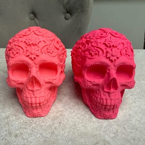 May include: Two vibrant pink decorative skull sculptures. Each skull features intricate floral and swirl patterns across the surface. The skulls are displayed on a light gray surface, showcasing their detailed design.