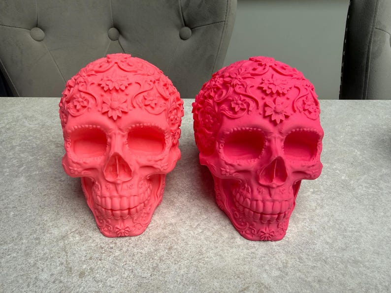 May include: Two vibrant pink decorative skull sculptures. Each skull features intricate floral and swirl patterns across the surface. The skulls are displayed on a light gray surface, showcasing their detailed design.