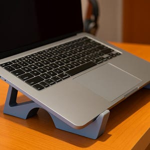 May include: A silver laptop computer is placed on a gray laptop stand. The laptop has a black keyboard and a black screen. The stand is angled, and the laptop is positioned on a wooden desk.