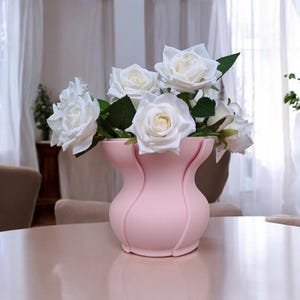 Puede incluir: Un jarr&oacute;n rosa con un dise&ntilde;o curvo &uacute;nico contiene un ramo de rosas blancas con hojas verdes. El jarr&oacute;n est&aacute; sobre una mesa rosa claro, creando una est&eacute;tica suave y elegante. Las rosas est&aacute;n en plena floraci&oacute;n.