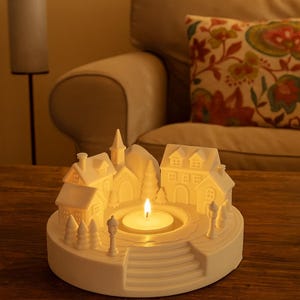 May include: A white ceramic candle holder shaped like a village scene, featuring miniature houses, a church, and trees. A lit candle sits in the center, casting a warm glow. The holder is on a wooden surface.