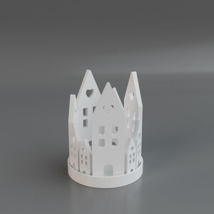 May include: A white, cylindrical candle holder with a cut-out village design. The holder features various house shapes with windows and heart-shaped cutouts. The design is repeated around the circumference of the holder.