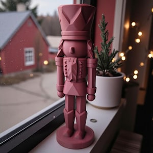 May include: A dusty rose-colored nutcracker figurine with a geometric design. The decorative piece is standing on a windowsill, with a small potted evergreen tree and a red building visible in the background. The nutcracker is a classic holiday decoration.