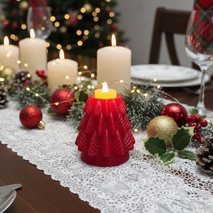 May include: A Christmas table setting featuring a red tree-shaped candle. The table is adorned with lit candles, red baubles, pine cones, and a white lace runner. A Christmas tree is visible in the background, creating a festive atmosphere.