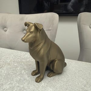 May include: A gold-colored dog statue in a seated position. The sculpture has a smooth finish and detailed features, including the dog's ears, snout, and paws. The statue is placed on a textured, light-colored surface.