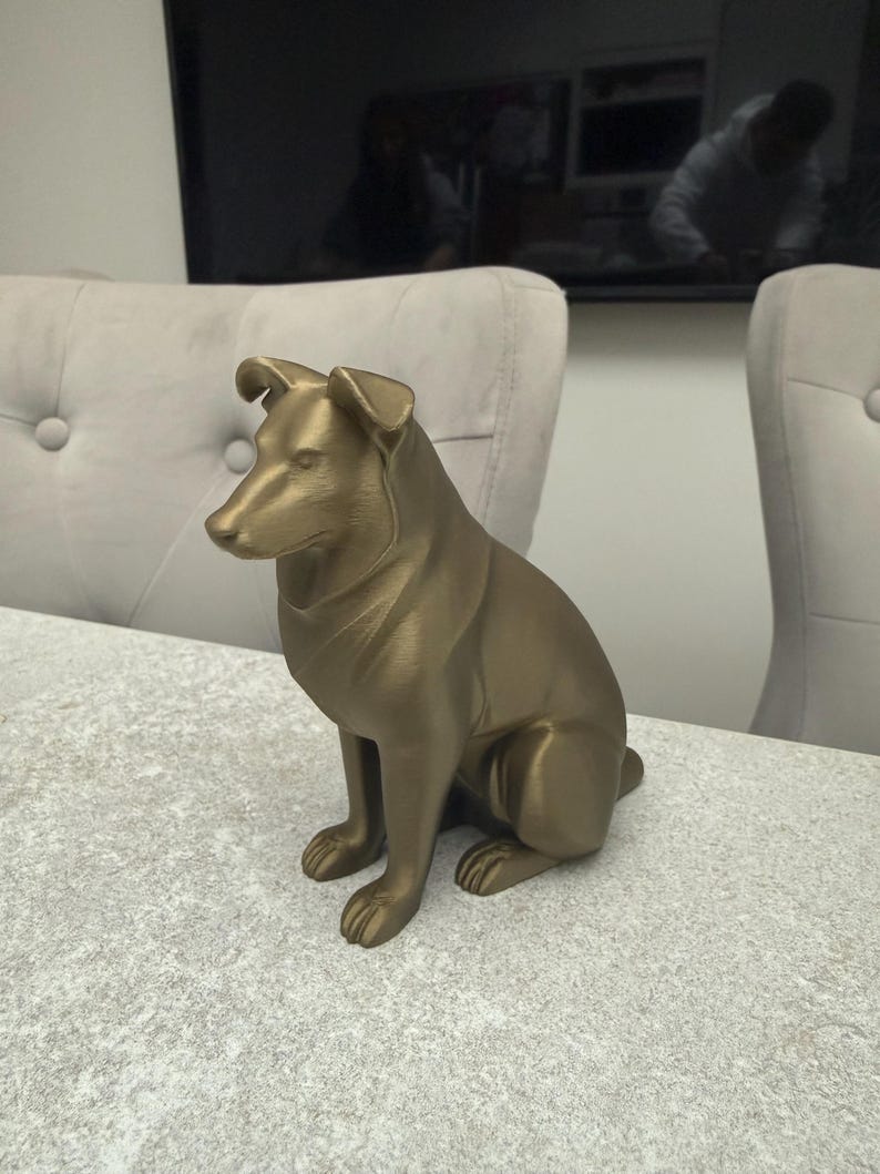 May include: A gold-colored dog statue in a seated position. The sculpture has a smooth finish and detailed features, including the dog's ears, snout, and paws. The statue is placed on a textured, light-colored surface.
