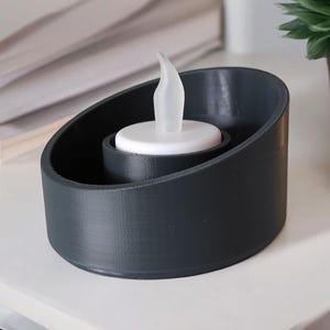 May include: A dark gray candle holder with a white LED candle. The holder has a rounded, asymmetrical design. The candle has a flame-shaped tip. A stack of white papers and a small potted plant are in the background.