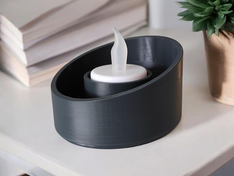 May include: A dark gray candle holder with a white LED candle. The holder has a rounded, asymmetrical design. The candle has a flame-shaped tip. A stack of white papers and a small potted plant are in the background.