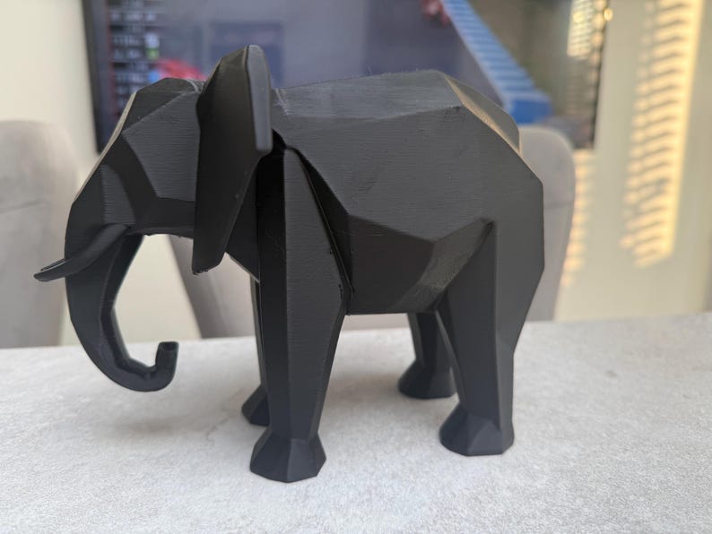 Op de afbeelding: Een zwarte geometrische olifant sculptuur. De sculptuur is gemaakt van een matzwart materiaal en heeft een facetontwerp. De olifant heeft grote oren, een slurf en vier poten. Dit decoratieve item is geschikt voor woondecoratie.