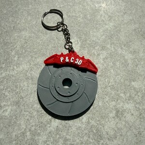 May include: A gray 3D-printed keychain featuring a brake rotor design with a red brake caliper. The caliper has the text "P & C 3D" printed on it. The keychain includes a silver key ring and chain.