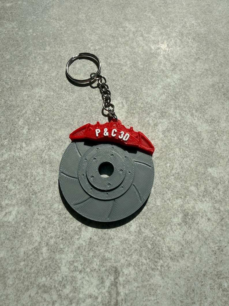 May include: A gray 3D-printed keychain featuring a brake rotor design with a red brake caliper. The caliper has the text "P & C 3D" printed on it. The keychain includes a silver key ring and chain.