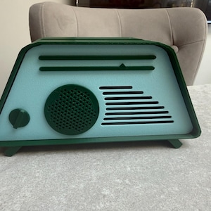 May include: A retro-style radio in shades of teal and green. The radio features a circular speaker, horizontal lines, and a tuning dial. The design is reminiscent of vintage electronics, with a modern color palette.