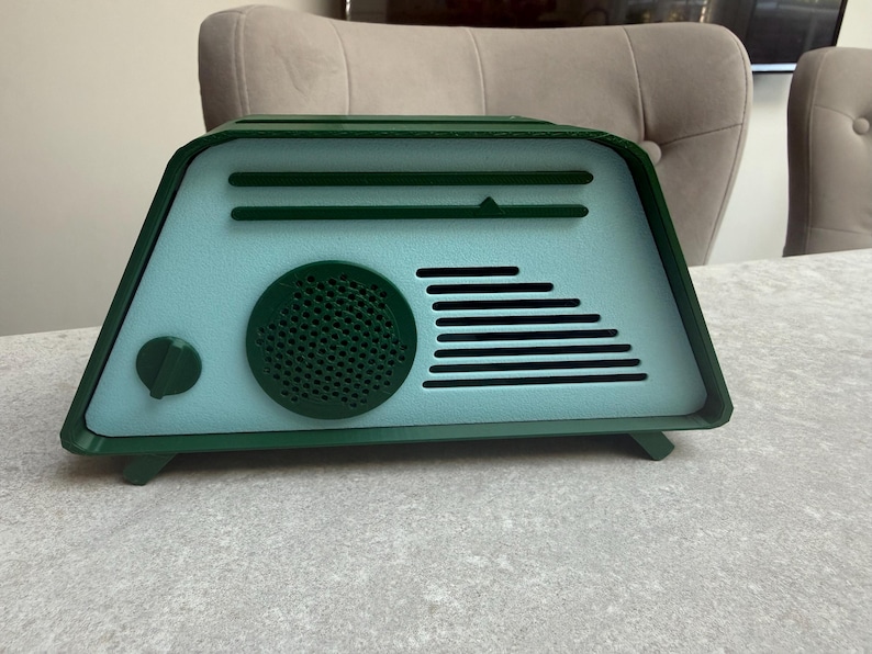 May include: A retro-style radio in shades of teal and green. The radio features a circular speaker, horizontal lines, and a tuning dial. The design is reminiscent of vintage electronics, with a modern color palette.