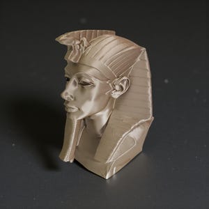 May include: A detailed bust of an Egyptian pharaoh, rendered in a metallic beige color. The sculpture features a stylized headdress and a serene facial expression, showcasing intricate details of the face and headwear. The bust is a decorative item.