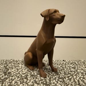 May include: A brown dog statue in a seated position. The statue has a smooth finish and is sitting on a patterned surface. The dog is looking to the right.