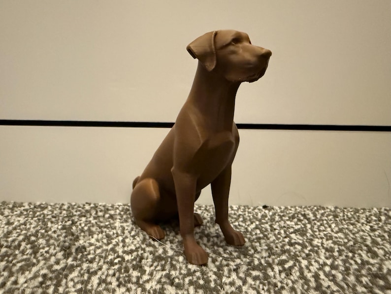 May include: A brown dog statue in a seated position. The statue has a smooth finish and is sitting on a patterned surface. The dog is looking to the right.