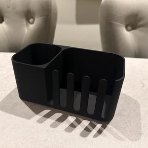 May include: A black, rectangular utensil holder with two compartments. One section is a solid square, and the other has vertical dividers. The holder is made of a matte material and sits on a light-colored surface.