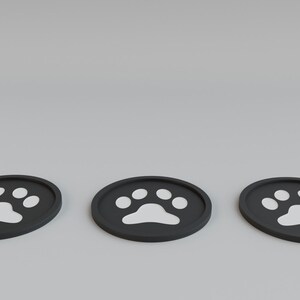 May include: Three black, oval-shaped coasters with a white paw print design. The coasters are arranged horizontally against a light gray background. These coasters are suitable for pet owners.