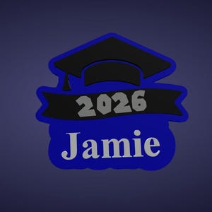 K&ouml;nnte beinhalten: Ein blau-schwarzes Design zum Thema Abschluss mit dem Namen "Jamie" und dem Jahr "2026". Das Design beinhaltet eine Abschlusskappe und ein Banner. Der Text ist in einer Kontrastfarbe gehalten, wodurch er sich vom blauen Hintergrund abhebt.