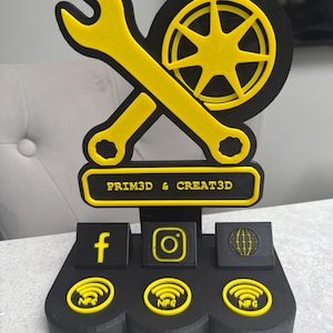 May include: A black and yellow 3D-printed desk organizer featuring a wrench and wheel design. The organizer includes social media icons for Facebook, Instagram, and a globe, along with the text "PRIM3D & CREAT3D". It also has NFC symbols.