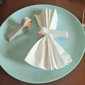 May include: A light blue plate with a white napkin folded into a fan shape, and a bird-shaped napkin ring. A second bird-shaped napkin ring is on the plate. A fork and knife are on the right side of the plate.