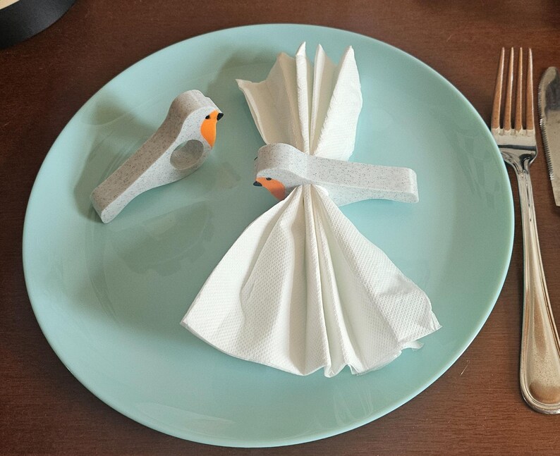 May include: A light blue plate with a white napkin folded into a fan shape, and a bird-shaped napkin ring. A second bird-shaped napkin ring is on the plate. A fork and knife are on the right side of the plate.