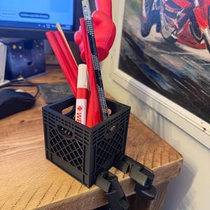 May include: A black, miniature crate-shaped desk organizer with attached clips, holding various red and black writing utensils. A red foam hand and a pencil with a dotted pattern are also visible. The organizer sits on a wooden surface.