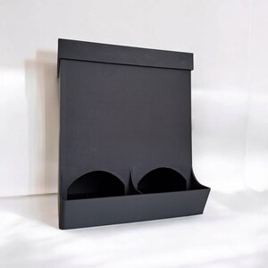 May include: A matte black wall-mounted organizer with two circular openings and a lower shelf. The organizer has a rectangular shape with a flat top and is designed to hold small items.