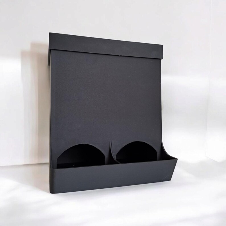 May include: A matte black wall-mounted organizer with two circular openings and a lower shelf. The organizer has a rectangular shape with a flat top and is designed to hold small items.