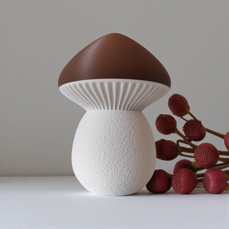 May include: A ceramic mushroom-shaped decorative item with a brown cap and a white textured stem. The mushroom has a ribbed underside and sits next to a sprig of red berries. The overall design is whimsical and nature-inspired.