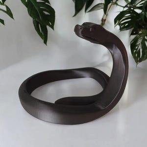 Puede incluir: Una escultura de serpiente marr&oacute;n oscuro con un cuerpo curvado y la cabeza levantada. La escultura est&aacute; hecha de un material liso y se encuentra sobre una superficie blanca. Se ven hojas verdes en el fondo.